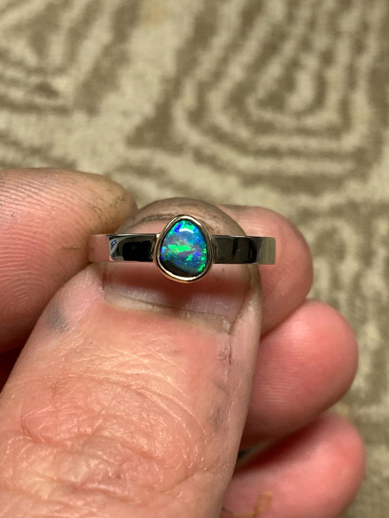 May include: A silver ring with a pear-shaped opal gemstone. The opal displays a vibrant play of color, with flashes of blue and green. The ring has a simple, modern design, showcasing the gemstone's natural beauty.