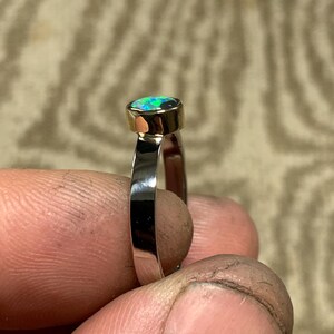 May include: A silver ring with a gold bezel setting an oval-shaped opal gemstone. The opal displays a vibrant play of color, with flashes of blue and green. The ring is held up by a person's fingers.