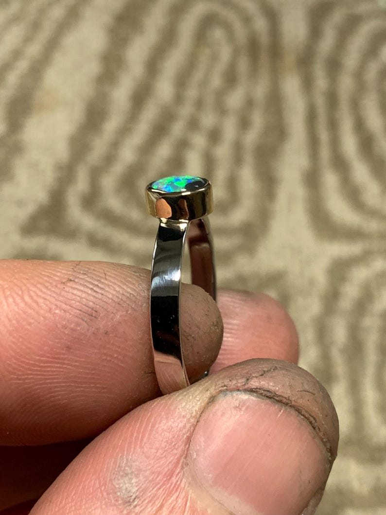 May include: A silver ring with a gold bezel setting an oval-shaped opal gemstone. The opal displays a vibrant play of color, with flashes of blue and green. The ring is held up by a person's fingers.