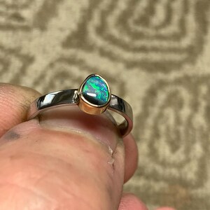 May include: A silver ring with a teardrop-shaped opal gemstone. The opal has a vibrant display of green and blue colors, set in a gold bezel. The ring is held between fingers, showcasing its polished band.