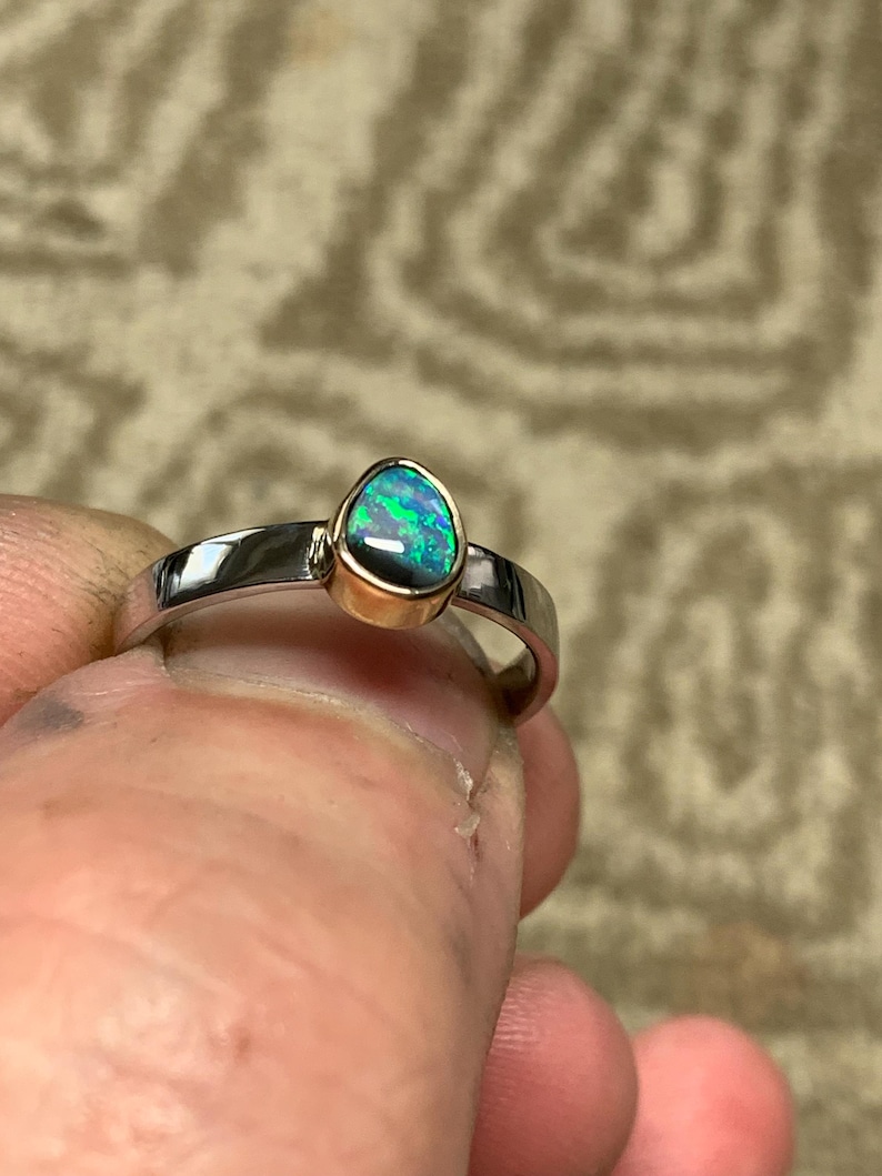 May include: A silver ring with a teardrop-shaped opal gemstone. The opal has a vibrant display of green and blue colors, set in a gold bezel. The ring is held between fingers, showcasing its polished band.