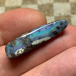 Könnte beinhalten: Ein polierter Opal-Edelstein in rechteckiger Form. Der Opal zeigt ein lebendiges Farbspiel mit Blau-, Grün- und Türkistönen. Der Stein ist mit dunkler Matrix und weißen Einschlüssen akzentuiert, was ein einzigartiges Muster erzeugt.