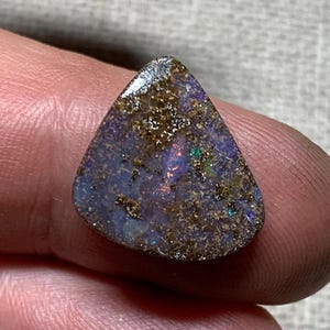 Könnte beinhalten: Ein dreieckiger Opal-Edelstein mit dunkelblauer Grundfarbe, der irisierende Blitze in Grün, Rot und Gold zeigt. Die Oberfläche hat ein strukturiertes Aussehen mit kleinen, goldbraunen Einschlüssen. Der Edelstein wird zwischen Fingern gehalten.