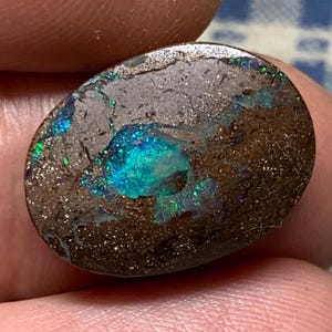 Könnte beinhalten: Ein ovaler Opal mit einer dunkelbraunen Basis und einem bläulich-grünen, irisierenden Schimmer. Der Opal hat eine raue, strukturierte Oberfläche.