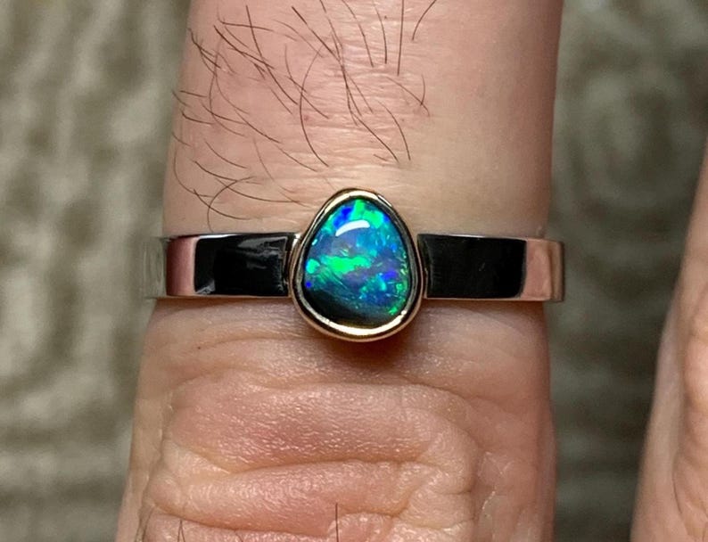 May include: A silver ring with a vibrant, pear-shaped opal gemstone. The opal displays a striking play of color, with flashes of blue and green. The gemstone is set in a gold bezel, adding a touch of elegance to the ring's design.