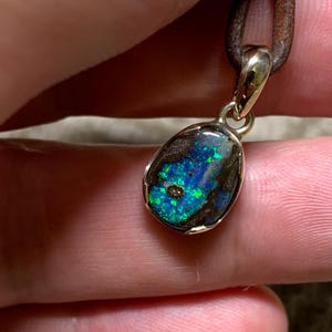 May include: A pendant with an opal gemstone in a gold setting, hanging from a brown leather cord. The opal exhibits a vivid play of colour, with blue and green flashes against a dark backdrop. This unique piece of jewellery would make a great gift.