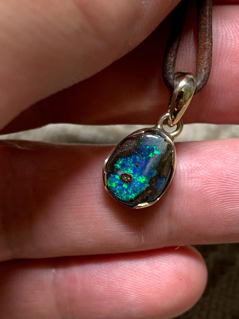 May include: A pendant with an opal gemstone in a gold setting, hanging from a brown leather cord. The opal exhibits a vivid play of colour, with blue and green flashes against a dark backdrop. This unique piece of jewellery would make a great gift.