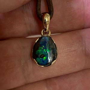 May include: A gold pendant featuring a dark opal stone. The opal exhibits striking green and blue colours within a dark base. The pendant hangs from a brown leather cord, presenting a natural and elegant design.