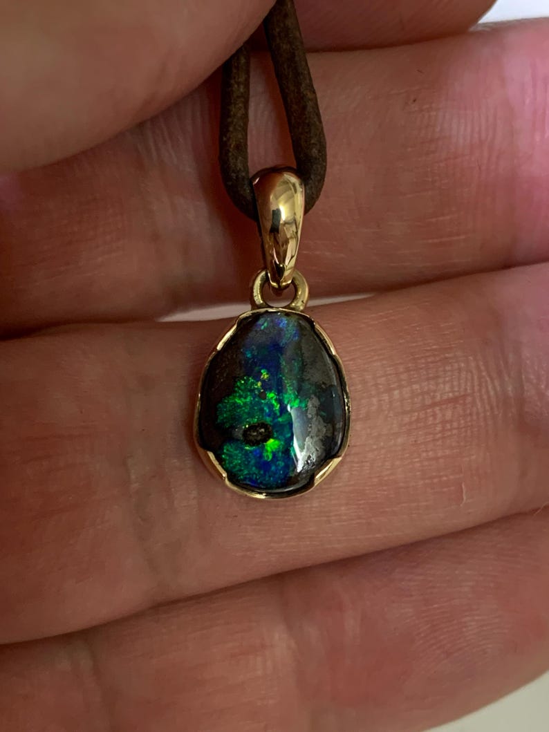 May include: A gold pendant featuring a dark opal stone. The opal exhibits striking green and blue colours within a dark base. The pendant hangs from a brown leather cord, presenting a natural and elegant design.