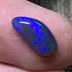 May include: A polished, oval opal gemstone. The opal displays a dark base colour with vibrant blue flashes. The stone is held between fingers, highlighting its natural beauty and unique colour play.