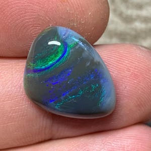 May include: A polished opal gemstone, teardrop-shaped. The stone has a dark grey base with internal flashes of green, blue, and purple. The surface is smooth and reflective, displaying a play of colour.
