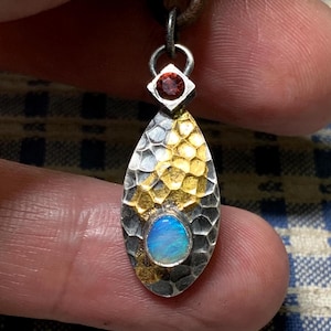 Può includere: Ciondolo fatto a mano con un design in argento, oro e opale. Il ciondolo presenta una forma a goccia in argento martellato con accenti dorati e un'opale ovale centrale. Una piccola gemma rossa è sopra.