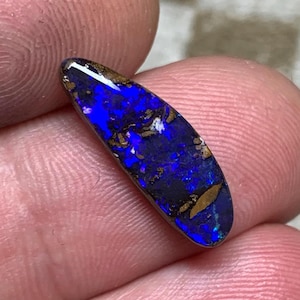 Könnte beinhalten: Ein polierter, oval geformter Opal-Edelstein. Der Opal zeigt eine lebendige blaue Farbe mit goldenen und braunen Reflexen. Der Edelstein wird zwischen zwei Fingern gehalten, was seine natürlichen Muster und Farben hervorhebt.