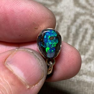 May include: A close-up of a small, oval opal pendant. The opal features vivid blue and green hues within a dark matrix, set in a silver or gold metal bezel. The pendant hangs from a delicate chain.