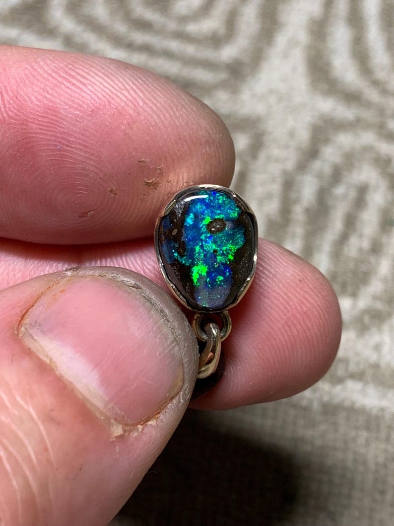 May include: A close-up of a small, oval opal pendant. The opal features vivid blue and green hues within a dark matrix, set in a silver or gold metal bezel. The pendant hangs from a delicate chain.
