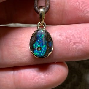 May include: A gold pendant featuring an oval opal gemstone. The opal showcases a vibrant play of colour, with flashes of blue and green. The pendant hangs from a brown leather cord, forming a simple yet elegant necklace.