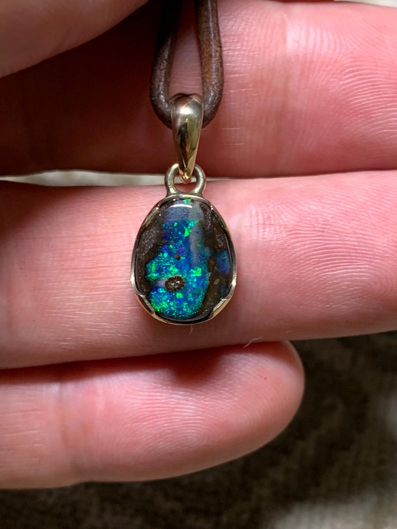 May include: A gold pendant featuring an oval opal gemstone. The opal showcases a vibrant play of colour, with flashes of blue and green. The pendant hangs from a brown leather cord, forming a simple yet elegant necklace.