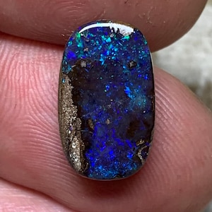 Könnte beinhalten: Ein polierter, ovaler Opal mit dunkler Grundfarbe und leuchtenden blauen und grünen Lichtreflexen. Der Edelstein hat eine strukturierte, goldbraune Matrix an einer Kante. Gehalten zwischen Fingern.