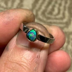 May include: A silver ring with a teardrop-shaped opal gemstone. The opal displays a vibrant play of color, with flashes of green and blue. The ring is held between fingers, showcasing the polished metal and the gemstone's unique shape.
