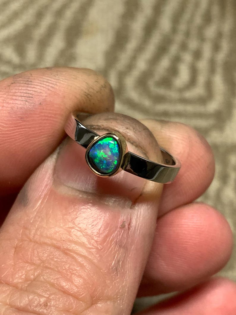 May include: A silver ring with a teardrop-shaped opal gemstone. The opal displays a vibrant play of color, with flashes of green and blue. The ring is held between fingers, showcasing the polished metal and the gemstone's unique shape.