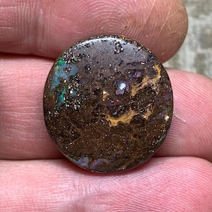 Könnte beinhalten: Ein runder, polierter Opal-Edelstein mit dunkelbrauner Grundfarbe. Der Opal zeigt schillernde Blitze in Grün, Blau und Gold auf seiner Oberfläche. Der Edelstein wird zwischen Fingern gehalten.