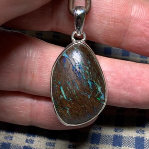 May include: A silver pendant featuring a brown and blue opal stone. The pendant is on a brown leather cord.