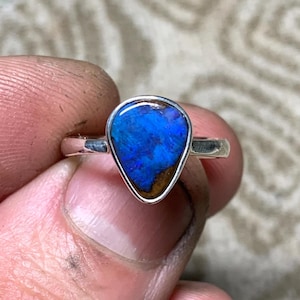 May include: A silver ring featuring a pear-shaped opal gemstone. The opal displays vibrant blue and brown hues, set within a silver bezel. The ring is held by a hand, showcasing the polished silver band and the gemstone's intricate color patterns.