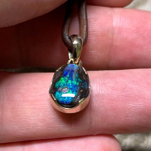 May include: A pendant with a colourful opal gemstone, displaying blue and green tones, set in a gold-coloured bezel. The opal is suspended from a brown leather cord. The pendant is held by a person's fingers.