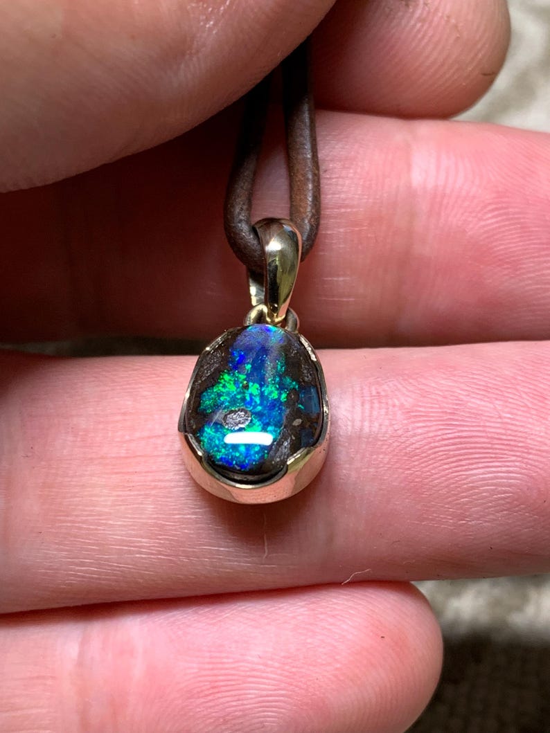 May include: A pendant with a colourful opal gemstone, displaying blue and green tones, set in a gold-coloured bezel. The opal is suspended from a brown leather cord. The pendant is held by a person's fingers.