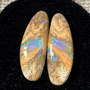 Peut inclure: Deux opales boulder allongées en forme de goutte avec une surface polie et lisse. Les pierres présentent un mélange de roche matricielle beige, sable et fauve, entremêlé d'éclats irisés vibrants de bleu, de vert et de violet.