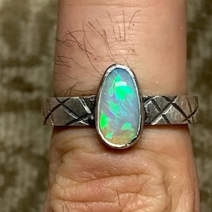 Op de afbeelding: Zilveren ring met een ovale opaal edelsteen met levendige groene en blauwe tinten. De band van de ring heeft een textuur met een kruispatroon. De opaal is gezet in een zilveren zetting, wat een opvallend contrast creëert.