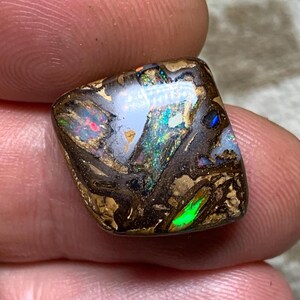 Könnte beinhalten: Ein polierter, quadratisch geschliffener Opal-Edelstein. Der Stein hat eine dunkelbraune Basis mit komplizierten Mustern aus Gold, Schwarz und schillernden Blitzen in Grün, Blau und Rot. Der Opal wird zwischen zwei Fingern gehalten.