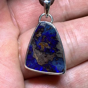Peut inclure: Un pendentif trapézoïdal composé d'une pierre d'opale boulder bleu foncé et marron sertie dans un cadre en argent poli. Le pendentif est fixé à un cordon en cuir marron par une bélière en argent, révélant un motif naturel de reflets bleu profond.