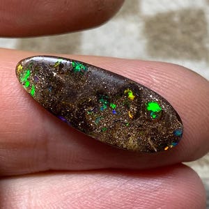 Könnte beinhalten: Ein ovaler, dunkelbrauner Boulder-Opal-Edelstein mit einer polierten Oberfläche, die lebhafte Blitze in leuchtendem Grün, Gelb und Blau aufweist, welche über die texturierte, erdige Matrix des Steins verteilt sind.