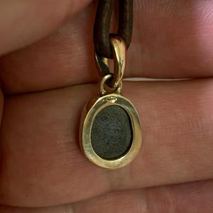 May include: A gold pendant with a dark grey stone set in an oval frame, suspended from a brown leather cord. The pendant's metal has a polished finish, and the cord appears to be securely attached. The design is simple and elegant.