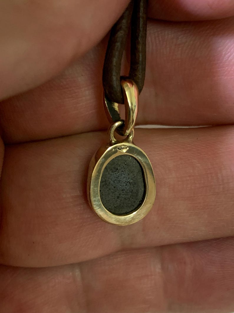 May include: A gold pendant with a dark grey stone set in an oval frame, suspended from a brown leather cord. The pendant's metal has a polished finish, and the cord appears to be securely attached. The design is simple and elegant.