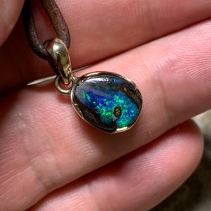 May include: A gold pendant featuring an opal stone with blue and green hues. The opal is set in a gold bezel and suspended from a brown leather cord. This pendant is a piece of jewellery.