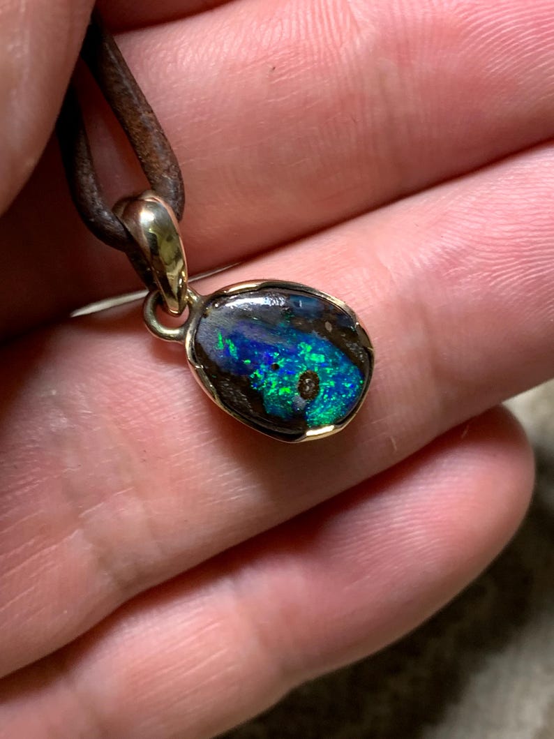 May include: A gold pendant featuring an opal stone with blue and green hues. The opal is set in a gold bezel and suspended from a brown leather cord. This pendant is a piece of jewellery.