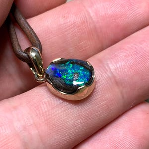 May include: A gold pendant featuring an oval opal gemstone. The opal showcases vivid blue and green hues, with a small diamond. The pendant hangs from a brown leather cord, creating a unique piece of jewellery.