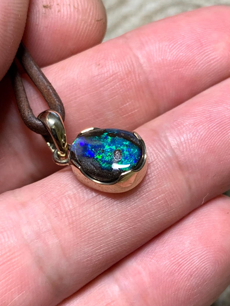 May include: A gold pendant featuring an oval opal gemstone. The opal showcases vivid blue and green hues, with a small diamond. The pendant hangs from a brown leather cord, creating a unique piece of jewellery.