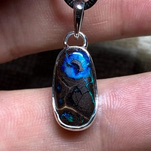 Puede incluir: Un colgante con un ópalo boulder de forma ovalada alargada engastado en un bisel de plata pulida. La piedra de color marrón oscuro muestra destellos iridiscentes y vibrantes de azul eléctrico y verde en su superficie, suspendido de un cordón negro.
