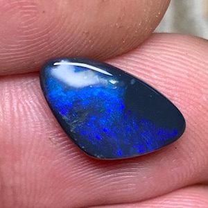 May include: A black opal gemstone with a polished surface, showcasing vivid blue and green colours. The opal is held between fingers and has a smooth, rounded shape. The gemstone displays a striking play of colour.