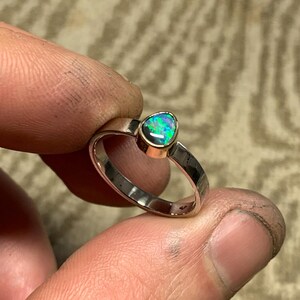 May include: A silver ring with a pear-shaped opal gemstone. The opal displays a vibrant play of color, with flashes of green, blue, and purple. The ring is held between fingers, showcasing the polished metal and gemstone.