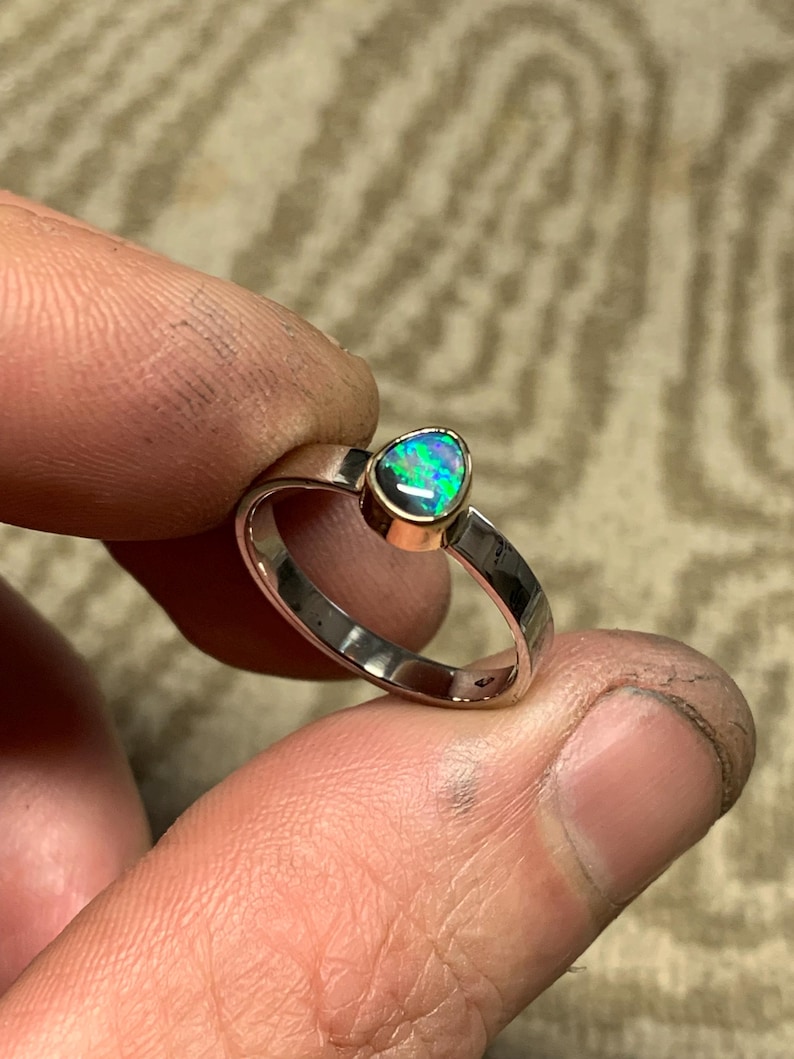 May include: A silver ring with a pear-shaped opal gemstone. The opal displays a vibrant play of color, with flashes of green, blue, and purple. The ring is held between fingers, showcasing the polished metal and gemstone.