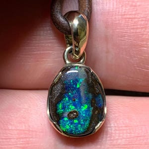 May include: An oval opal pendant with a gold-coloured metal frame, suspended from a brown leather cord. The opal displays a striking mix of blue and green hues. This unique pendant is a piece of jewellery with a distinctive colour pattern.