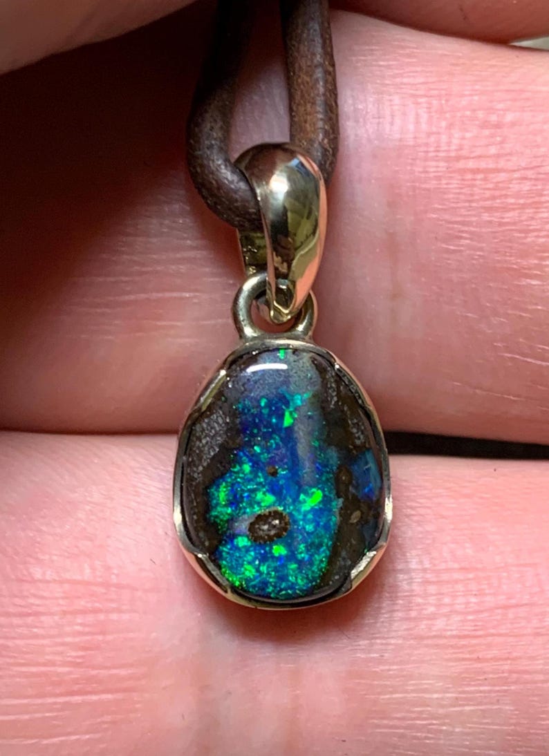 May include: An oval opal pendant with a gold-coloured metal frame, suspended from a brown leather cord. The opal displays a striking mix of blue and green hues. This unique pendant is a piece of jewellery with a distinctive colour pattern.