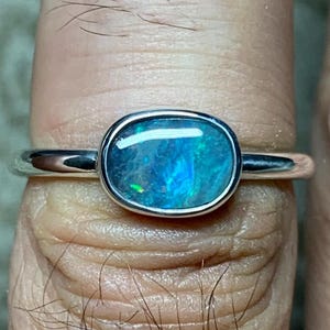 May include: A silver ring with an oval-shaped opal gemstone. The opal displays a vibrant play of color, with shades of blue, green, and hints of other colors. The ring's band is smooth and polished.
