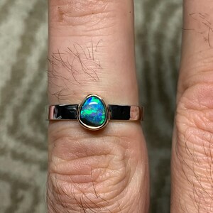 May include: A silver ring with a teardrop-shaped opal gemstone. The opal displays vibrant blue and green colors. The ring has a gold bezel setting and a polished silver band. A piece of jewelry.