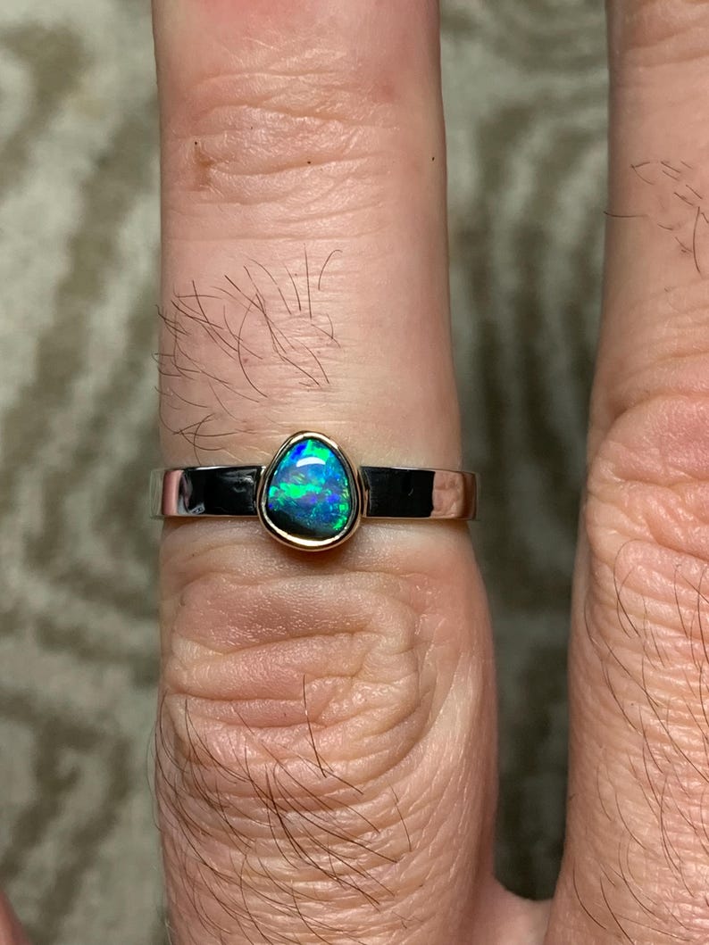 May include: A silver ring with a teardrop-shaped opal gemstone. The opal displays vibrant blue and green colors. The ring has a gold bezel setting and a polished silver band. A piece of jewelry.