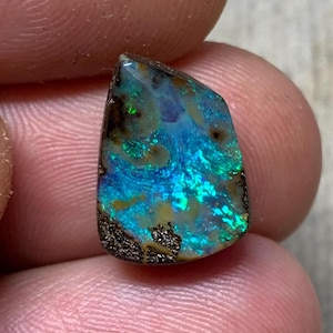 Peut inclure: Une pierre précieuse opale boulder polie de forme irrégulière présentant des éclats vibrants de bleu électrique et de vert sur une matrice de minerai de fer brun terreux avec des textures naturelles et des inclusions visibles.